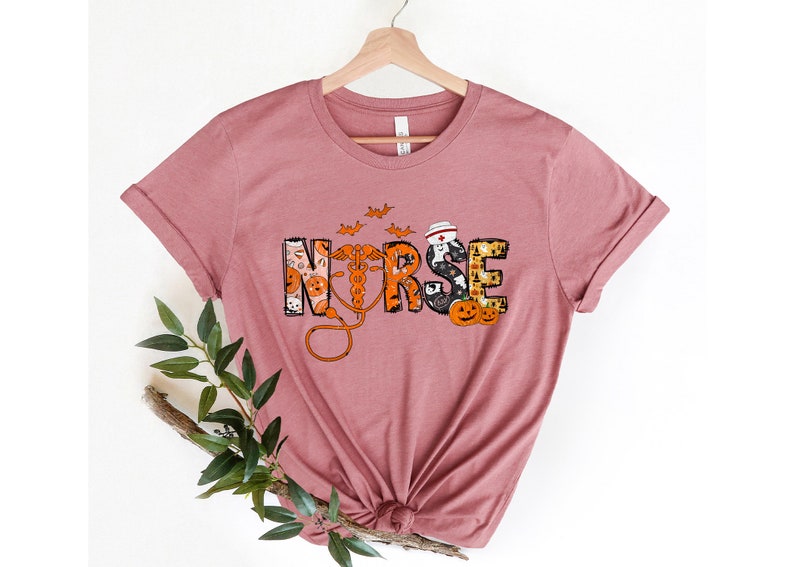 May include: Mauve t-shirt with the word "NURSE" in a Halloween-themed design. The letters are decorated with pumpkins, bats, and a medical caduceus. The shirt is displayed on a wooden hanger.