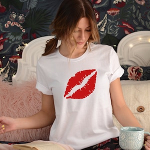 May include: White t-shirt with a red lipstick print. The shirt is worn by a person sitting on a bed with a pink fuzzy throw pillow.