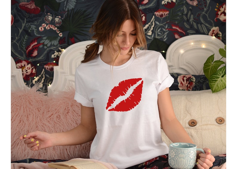 May include: White t-shirt with a red lipstick print. The shirt is worn by a person sitting on a bed with a pink fuzzy throw pillow.