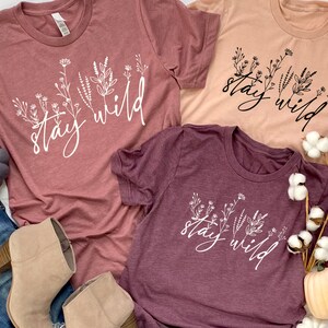 May include: Three t-shirts with the text "stay wild" in white lettering with a floral design. The shirts are in shades of pink and burgundy.