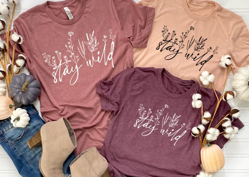 May include: Three t-shirts with the text "stay wild" in white lettering with a floral design. The shirts are in shades of pink and burgundy.