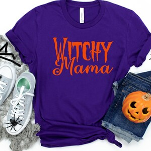 May include: A purple t-shirt with the words "Witchy Mama" in orange script. The shirt is paired with gray sneakers, denim shorts, and Halloween decorations, including spiders and a jack-o'-lantern.