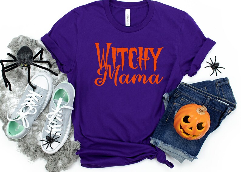 May include: A purple t-shirt with the words "Witchy Mama" in orange script. The shirt is paired with gray sneakers, denim shorts, and Halloween decorations, including spiders and a jack-o'-lantern.