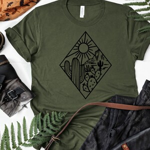K&ouml;nnte beinhalten: Ein gr&uuml;nes T-Shirt mit einer schwarzen Grafik einer W&uuml;stenlandschaft mit Kakteen und einer Sonne. Die Grafik ist in Form eines Diamanten.