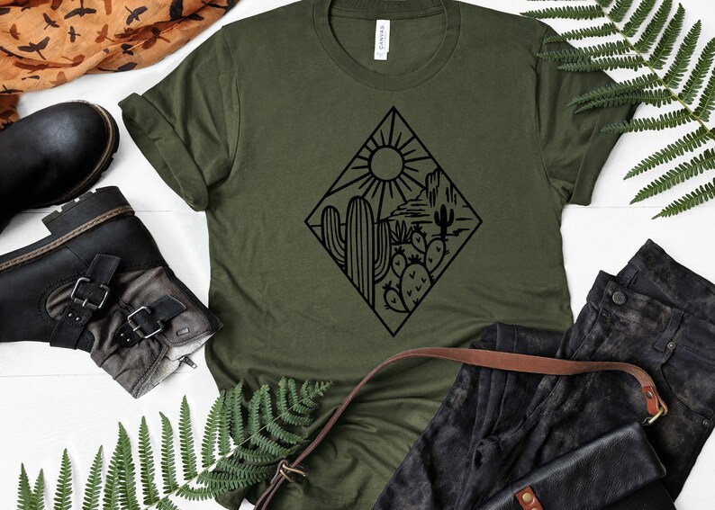 K&ouml;nnte beinhalten: Ein gr&uuml;nes T-Shirt mit einer schwarzen Grafik einer W&uuml;stenlandschaft mit Kakteen und einer Sonne. Die Grafik ist in Form eines Diamanten.