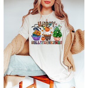 K&ouml;nnte beinhalten: Cremefarbenes T-Shirt mit einem farbenfrohen Aufdruck mit Cupcakes, die mit Halloween- und Thanksgiving-Themen dekoriert sind. Der Text "HAPPY HALLOTHANKSMAS" ist &uuml;ber und unter den Cupcakes zu sehen. Das Shirt ist eine festliche Mischung aus Feierlichkeiten.