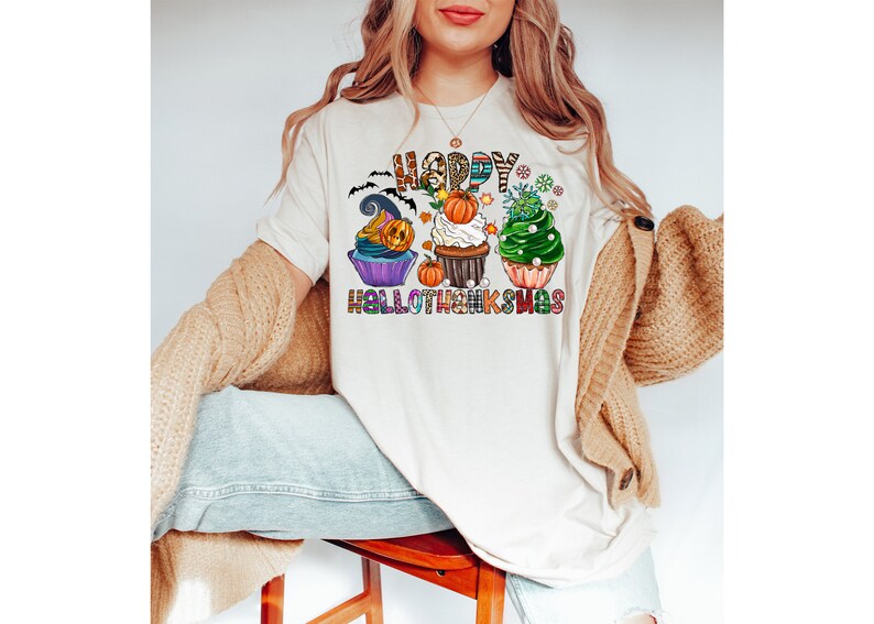 K&ouml;nnte beinhalten: Cremefarbenes T-Shirt mit einem farbenfrohen Aufdruck mit Cupcakes, die mit Halloween- und Thanksgiving-Themen dekoriert sind. Der Text "HAPPY HALLOTHANKSMAS" ist &uuml;ber und unter den Cupcakes zu sehen. Das Shirt ist eine festliche Mischung aus Feierlichkeiten.