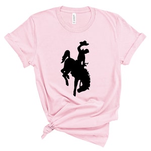 May include: A pink t-shirt with a black silhouette of a cowboy riding a bucking horse.