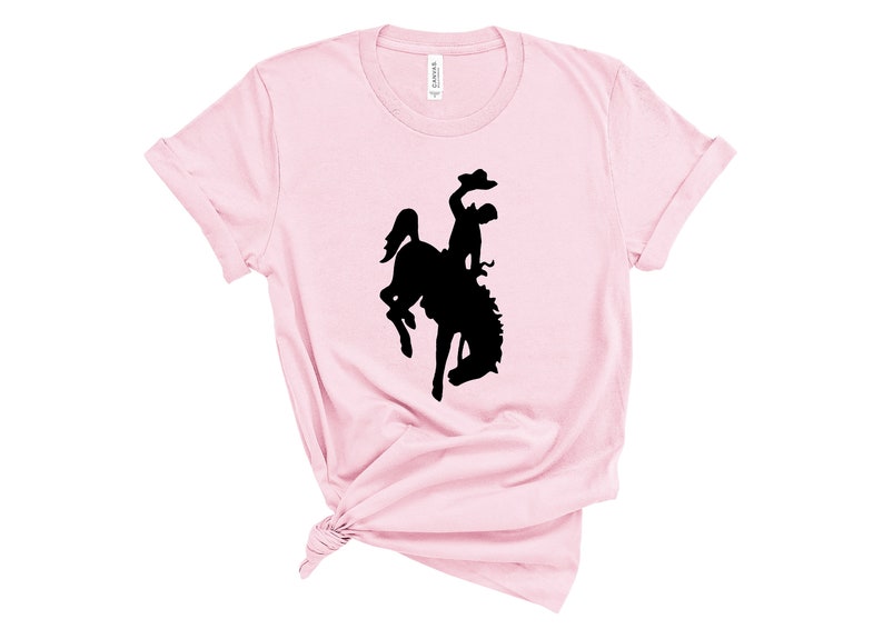 May include: A pink t-shirt with a black silhouette of a cowboy riding a bucking horse.