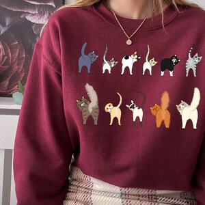 May include: A maroon sweatshirt with a graphic of multiple cats with their tails up in the air. The cats are all different colors and breeds.