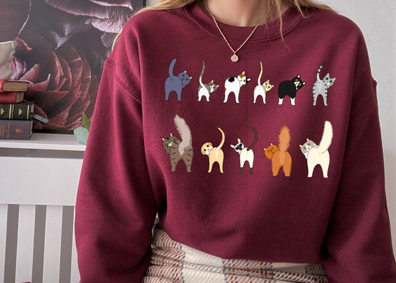 May include: A maroon sweatshirt with a graphic of multiple cats with their tails up in the air. The cats are all different colors and breeds.