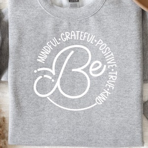 May include: Gray sweatshirt with a white circle design that says "Be" in a cursive font. The circle is surrounded by the words "Mindful, Grateful, Positive, True, Kind".
