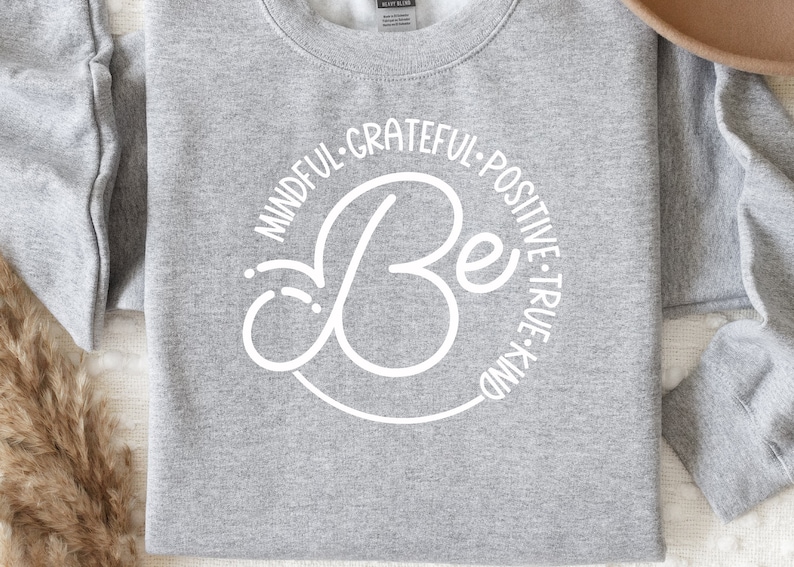 May include: Gray sweatshirt with a white circle design that says "Be" in a cursive font. The circle is surrounded by the words "Mindful, Grateful, Positive, True, Kind".
