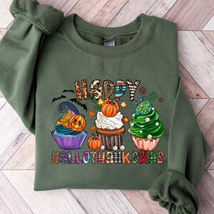 K&ouml;nnte beinhalten: Olivgr&uuml;nes Sweatshirt mit einem farbenfrohen Aufdruck mit der Aufschrift "HAPPY HALLOTHANKSMAS". Das Design zeigt drei Cupcakes, die mit Halloween-, Thanksgiving- und Weihnachtsmotiven dekoriert sind. Enth&auml;lt Flederm&auml;use, K&uuml;rbisse und Schneeflocken.