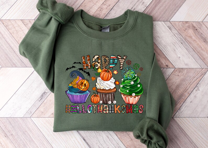 K&ouml;nnte beinhalten: Olivgr&uuml;nes Sweatshirt mit einem farbenfrohen Aufdruck mit der Aufschrift "HAPPY HALLOTHANKSMAS". Das Design zeigt drei Cupcakes, die mit Halloween-, Thanksgiving- und Weihnachtsmotiven dekoriert sind. Enth&auml;lt Flederm&auml;use, K&uuml;rbisse und Schneeflocken.