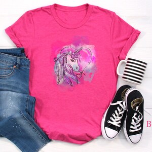 May include: Pink t-shirt with a watercolor unicorn design in a heart shape. The shirt is paired with blue jeans and black sneakers. "Berry Shirt" is written in pink text on the bottom right corner of the image.