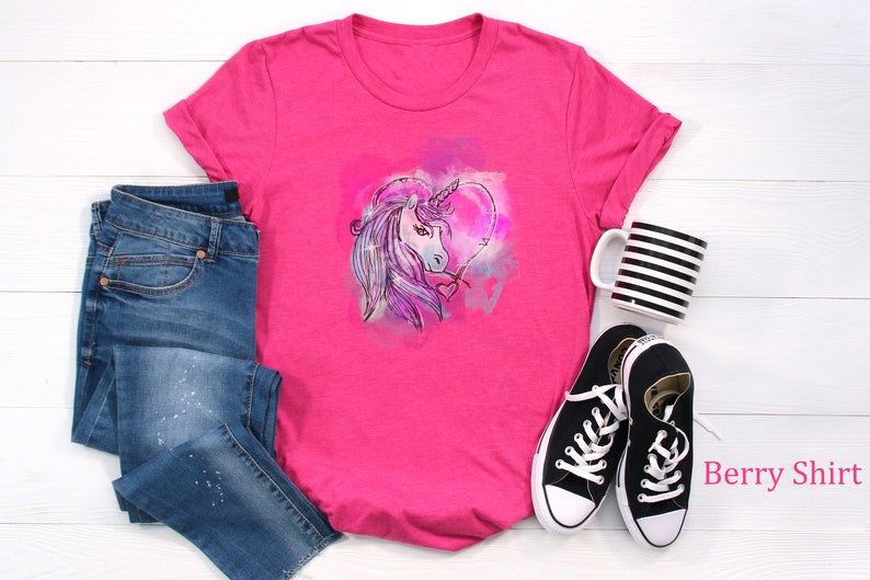 May include: Pink t-shirt with a watercolor unicorn design in a heart shape. The shirt is paired with blue jeans and black sneakers. "Berry Shirt" is written in pink text on the bottom right corner of the image.