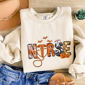 May include: Cream-colored sweatshirt with the word "NURSE" in a Halloween-themed design. The design includes pumpkins, bats, and a stethoscope. The sweatshirt is paired with blue jeans, a hat, and sandals.