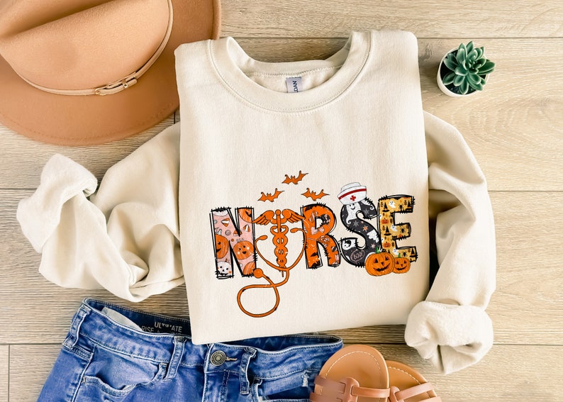 May include: Cream-colored sweatshirt with the word "NURSE" in a Halloween-themed design. The design includes pumpkins, bats, and a stethoscope. The sweatshirt is paired with blue jeans, a hat, and sandals.