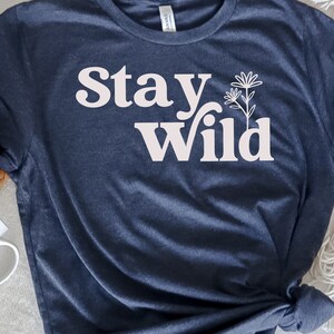 May include: A navy blue t-shirt with the text "Stay Wild" in white lettering. The text is in a retro font style. There is a small white flower graphic next to the text.
