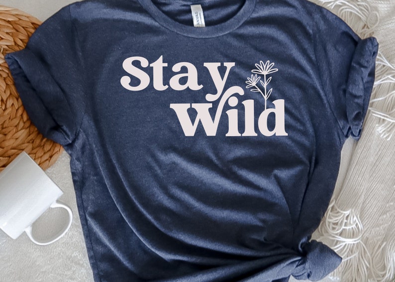May include: A navy blue t-shirt with the text "Stay Wild" in white lettering. The text is in a retro font style. There is a small white flower graphic next to the text.
