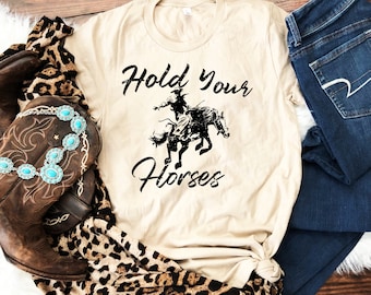 May include: A beige t-shirt with a black graphic of a cowboy riding a bucking horse. The text "Hold Your Horses" is printed below the graphic.