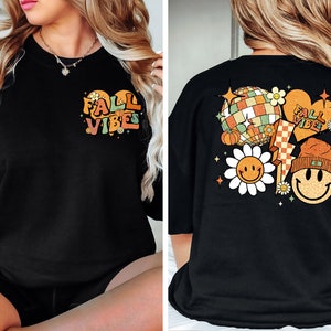 May include: Black oversized t-shirt with a graphic print of a heart with the text "Fall Vibes" on the front and a graphic print of a disco ball, pumpkin, lightning bolt, and two smiley faces with flowers on the back.