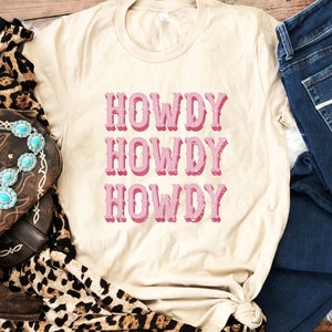 Op de afbeelding: Een beige T-shirt met de tekst "Howdy Howdy Howdy" in roze op de voorkant. Het shirt is gestyled met een bruine cowboylaars, een sjaal met luipaardprint en een blauwe jeans.