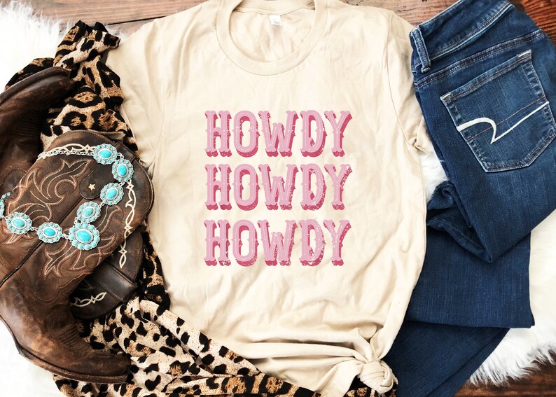 Op de afbeelding: Een beige T-shirt met de tekst "Howdy Howdy Howdy" in roze op de voorkant. Het shirt is gestyled met een bruine cowboylaars, een sjaal met luipaardprint en een blauwe jeans.