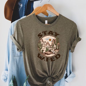 May include: A green t-shirt with a brown graphic of a desert landscape with cacti and the text "STAY WILD" in a circle.