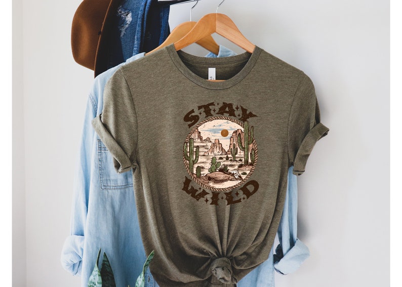 May include: A green t-shirt with a brown graphic of a desert landscape with cacti and the text "STAY WILD" in a circle.