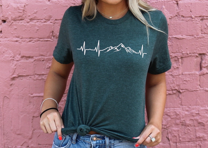 K&ouml;nnte beinhalten: Ein dunkelgr&uuml;nes T-Shirt mit einem wei&szlig;en Bergketten-Design und einer Herzschlaggrafik. Das Shirt ist aus einem weichen, bequemen Material gefertigt.