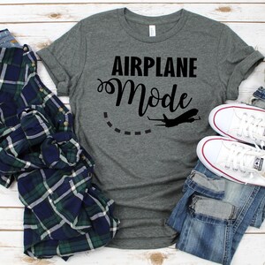 May include: A heather gray t-shirt with the text "Airplane Mode" in black lettering. A small airplane silhouette is above the text. The shirt is laid out on a white wood background with a plaid shirt, jeans, and a small white pumpkin.