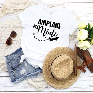 May include: White t-shirt with the text "Airplane Mode" in black lettering, a dashed line, and a small airplane graphic. The shirt is laid out on a white wooden surface with a straw hat, denim shorts, sandals, and a white lace shawl.