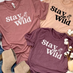 May include: Three t-shirts with the text "Stay Wild" printed on them. The shirts are in different colors: pink, peach, and burgundy. Each shirt has a small flower graphic next to the text.