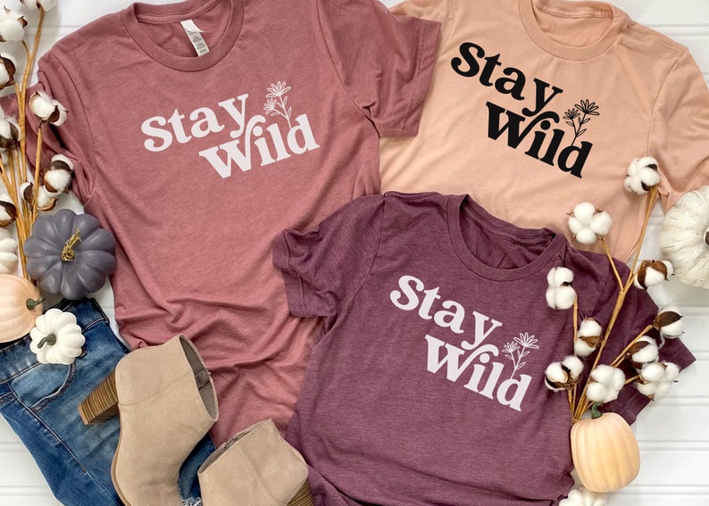 May include: Three t-shirts with the text "Stay Wild" printed on them. The shirts are in different colors: pink, peach, and burgundy. Each shirt has a small flower graphic next to the text.
