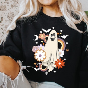 May include: Black sweatshirt with a retro ghost design. The illustration includes a smiling ghost, flowers, bats, a rainbow, and stars. The design has a vintage aesthetic, suitable for Halloween or casual wear.