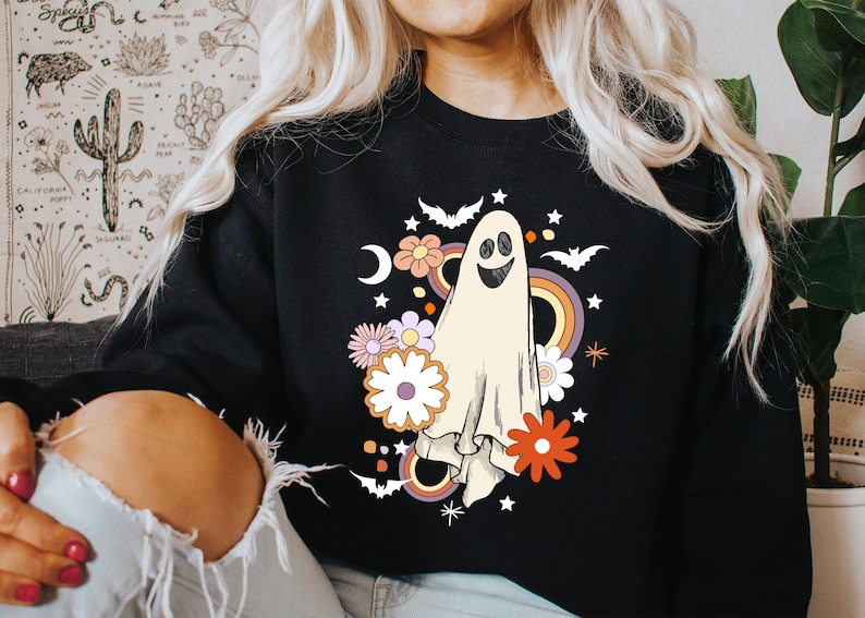 May include: Black sweatshirt with a retro ghost design. The illustration includes a smiling ghost, flowers, bats, a rainbow, and stars. The design has a vintage aesthetic, suitable for Halloween or casual wear.