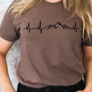 K&ouml;nnte beinhalten: Ein braunes T-Shirt mit einem schwarzen Bergpanorama und einem Herzschlag-Motiv. Die Grafik zeigt ein stilisiertes Bergpanorama mit einer Herzschlaglinie, die durch das Motiv verl&auml;uft.