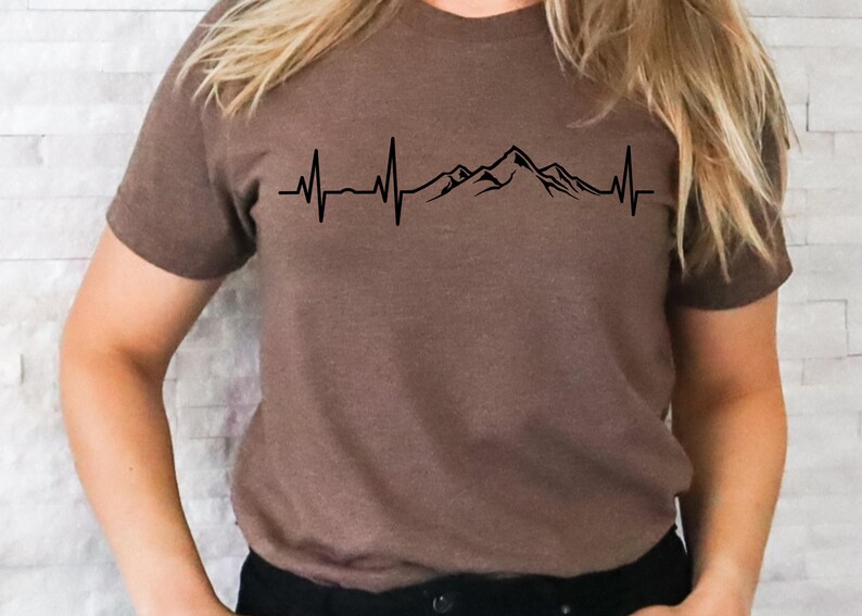 K&ouml;nnte beinhalten: Ein braunes T-Shirt mit einem schwarzen Bergpanorama und einem Herzschlag-Motiv. Die Grafik zeigt ein stilisiertes Bergpanorama mit einer Herzschlaglinie, die durch das Motiv verl&auml;uft.