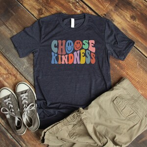 May include: A navy blue t-shirt with the text "CHOOSE KINDNESS" in rainbow colors with smiley faces. The shirt is laid out on a wooden surface with a pair of gray sneakers and khaki cargo pants.