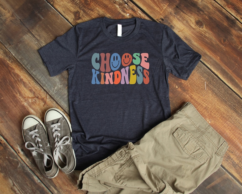 May include: A navy blue t-shirt with the text "CHOOSE KINDNESS" in rainbow colors with smiley faces. The shirt is laid out on a wooden surface with a pair of gray sneakers and khaki cargo pants.