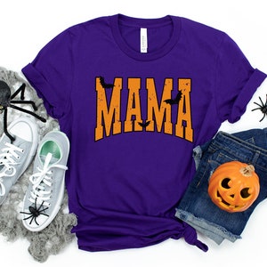 May include: A purple t-shirt with the word "MAMA" in orange, decorated with black bats. The shirt is paired with denim shorts, gray sneakers, and Halloween decorations, including spiders and a jack-o'-lantern.