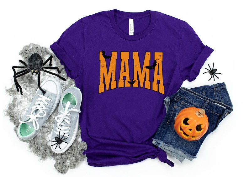 May include: A purple t-shirt with the word "MAMA" in orange, decorated with black bats. The shirt is paired with denim shorts, gray sneakers, and Halloween decorations, including spiders and a jack-o'-lantern.