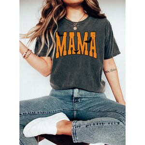 May include: Dark gray t-shirt with the word "MAMA" in orange block letters, with bat silhouettes. The shirt is worn with blue jeans and white sneakers. The overall theme is Halloween.