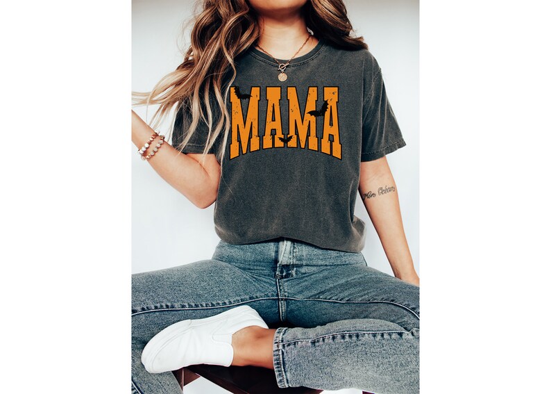 May include: Dark gray t-shirt with the word "MAMA" in orange block letters, with bat silhouettes. The shirt is worn with blue jeans and white sneakers. The overall theme is Halloween.
