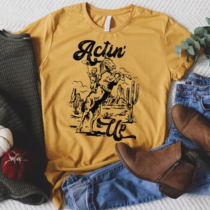 May include: A mustard yellow t-shirt with a black graphic of a cowboy riding a bucking horse. The text "Actin' Up" is printed in black below the graphic.