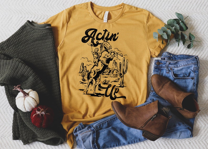 May include: A mustard yellow t-shirt with a black graphic of a cowboy riding a bucking horse. The text "Actin' Up" is printed in black below the graphic.