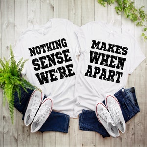 Op de afbeelding: Twee witte T-shirts met zwarte tekst. Het eerste shirt zegt "Nothing Makes Sense When We're" en het tweede shirt zegt "Apart".
