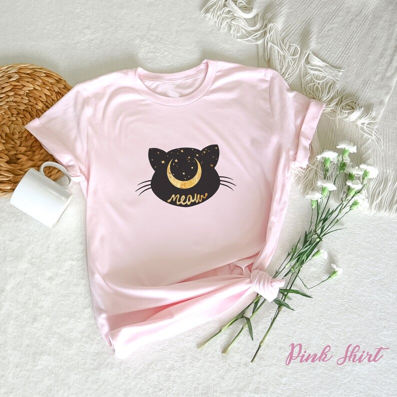 May include: A pink t-shirt with a black cat silhouette and a gold crescent moon with stars. The text "meow" is written in gold below the cat.